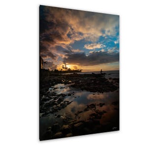 May include: A scenic photograph captures a sunset over a rocky shoreline. The sky is filled with dramatic clouds in shades of orange, yellow, and blue. A person stands on the rocks, silhouetted against the horizon. The water reflects the sky's colors.