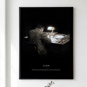 May include: A black and white framed wall art print featuring a person sitting at a desk illuminated by a desk lamp. The text FLOW is printed above the quote: The hours you don't feel passing are the ones that count. The frame is black.
