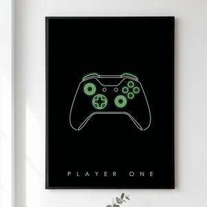 May include: A black framed wall art print featuring a white line drawing of a video game controller with glowing light green accents on the buttons and directional pad. The text PLAYER ONE is printed in white capital letters at the bottom centre.