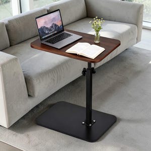May include: A dark brown, adjustable laptop table with a black base and support. A laptop, an open book, and a small vase of flowers sit on the table. The table is positioned next to a gray sofa on a gray rug.