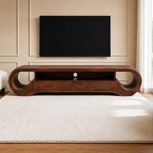 Puede incluir: Un mueble de televisión de madera marrón oscuro con un diseño largo y bajo, con lados curvos y abiertos. La unidad incluye un estante central abierto con un recorte circular para cables y tres cajones horizontales debajo del estante.
