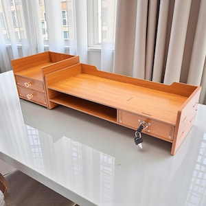 May include: A wooden desk organizer with a light brown finish. It features a monitor stand, two drawers, and a small cabinet with a lock and key. The organizer is designed to hold office supplies and electronics.