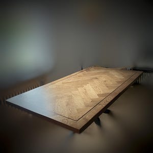 Op de afbeelding: Een rechthoekige houten tafel met een visgraatpatroon op het tafelblad. De tafel heeft een donkerbruine rand en poten. Het hout heeft een natuurlijke, lichtbruine kleur.