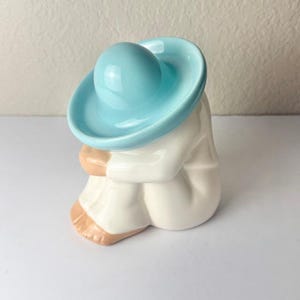 May include: A ceramic figurine of a seated figure wearing a light blue sombrero and a white robe. The figure is depicted in a crouching position with visible skin-toned feet and hands tucked near the face, finished with a smooth, glossy glaze.