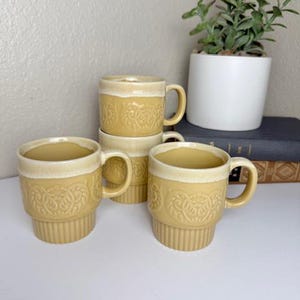 May include: Four stacked ceramic mugs with a yellow-tan color and a cream-colored rim. The mugs have a ribbed base and a decorative embossed floral pattern. A small plant in a white pot sits in the background.