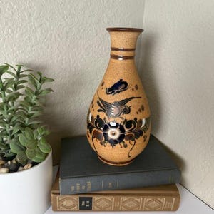 May include: A tan ceramic vase with a narrow neck and a wide base. The vase is decorated with black, blue, and white floral and bird designs. The vase is sitting on top of two books.