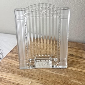May include: A clear glass picture frame featuring a vertical striped pattern on the central panel, flanked by two smooth, pillar-like side columns. The top and bottom sections are decorated with a fan-shaped, sunburst-style etched design.