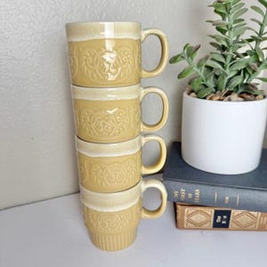 May include: A stack of four yellow ceramic mugs with a cream-colored rim and handle. Each mug features a detailed embossed floral design. The mugs are stacked on a white surface, next to a small potted plant and books.