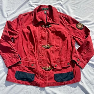 May include: A red jacket featuring a collar, long sleeves, and three brass-toned metal toggle closures down the front. The garment includes two lower pockets with dark blue flaps and a circular embroidered patch on the left sleeve.
