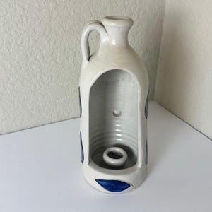 May include: A cream-colored ceramic candle holder shaped like a jug with a handle at the top. The front features a large arched cutout revealing a central candle cup and a small circular hole in the back wall. Dark blue accents decorate the exterior.
