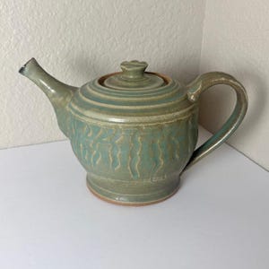 May include: A sage green ceramic teapot featuring a rounded body with vertical textured indentations, a matching lid with a small knob handle, a curved spout, and a sturdy side handle. The glaze has subtle variations in tone and texture.