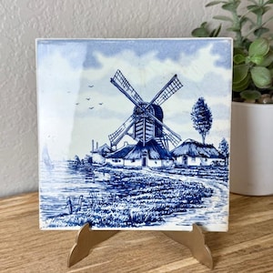 May include: A square ceramic tile featuring a blue and white transferware illustration of a traditional Dutch landscape. The scene depicts a large windmill, thatched-roof cottages, a calm body of water, and birds flying in a cloudy sky.