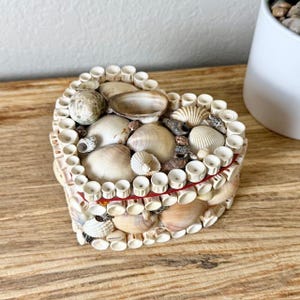 May include: A heart-shaped box decorated with various white, tan, and brown seashells. The lid and sides are covered in a mosaic of shells and small white cylindrical pieces, with a thin red line visible along the edge of the lid.