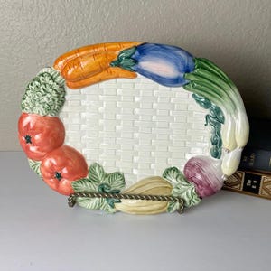 May include: An oval ceramic serving platter featuring a raised, embossed basket-weave pattern in the center. The rim is decorated with a colorful, three-dimensional arrangement of painted vegetables, including carrots, eggplant, tomatoes, and onions.