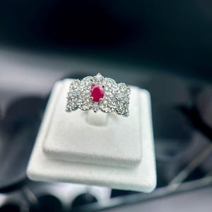Puede incluir: Un anillo de plata con una gema central de rubí ovalada, rodeada de gemas claras más pequeñas. El anillo se exhibe sobre una superficie de terciopelo blanco sobre un fondo oscuro. El diseño presenta intrincados filigranas y una forma de corona.