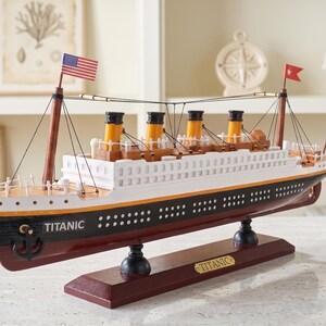 May include: A wooden model of the Titanic ship, featuring a white hull with black and yellow accents. The model has four smokestacks, multiple flags, and the name "Titanic" on the bow. It sits on a dark brown base.