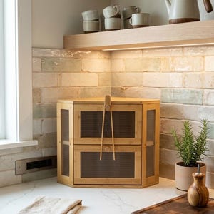 May include: A two-tiered, light brown bamboo cabinet with a pair of tongs hanging from the top. The cabinet has a slatted design on the doors. A folded beige linen towel sits on the countertop. A small potted plant and a wooden cutting board are also visible.