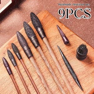 May include: A set of 9PCS handmade textured pens featuring wooden handles and black textured tips of varying sizes. The collection includes several long-handled tools with pointed ends, a small conical cap, and a circular brush-like attachment.
