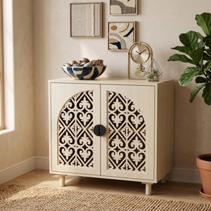May include: A light-colored wooden cabinet featuring two doors with arched, intricate cut-out patterns and black circular handles. The cabinet stands on four short legs and is topped with a decorative bowl filled with various stones and spheres.