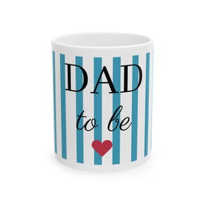 Puede incluir: Taza de cerámica blanca con rayas azules verticales y las palabras "DAD to be" en escritura negra. Un corazón rojo está centrado debajo del texto. La taza es de tamaño estándar para café o té.