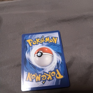 Peut inclure: Une carte à collectionner Pokémon bleue avec le logo Pokémon et un design de Poké Ball. La carte présente une Poké Ball blanche et rouge au centre, entourée d'un fond bleu tourbillonnant. Le mot "Pokémon" est imprimé en jaune.