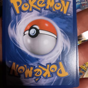 Puede incluir: Dorso de una carta coleccionable de Pokémon con fondo azul y la palabra "Pokémon" en amarillo. Una Poké Ball está centrada, con colores rojo, blanco y negro. La carta tiene un acabado brillante.