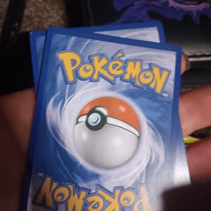 May include: The backs of three Pokémon trading cards are visible, featuring a blue background with the Pokémon logo in yellow and a Poké Ball design. The cards are held in a hand.