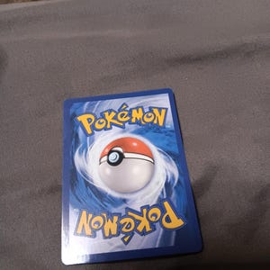 May include: A rectangular trading card featuring the blue and yellow Pokémon logo at the top and bottom. The center displays a circular red and white Poké Ball graphic surrounded by a swirling blue and white vortex design on a dark blue background.