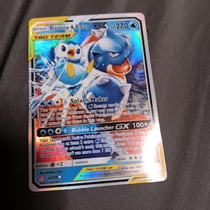 May include: A holographic Pokémon trading card featuring Blastoise & Piplup GX. The card has a blue and white color scheme with the text "TAG TEAM" and "Splash Maker". The card is a collectible item.