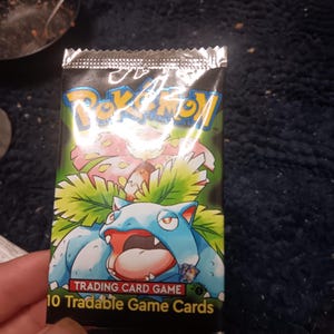 May include: Sealed Pokémon trading card pack featuring a blue and green Venusaur illustration. The pack has the Pokémon logo and text that reads "10 Tradable Game Cards". The background is black and green.