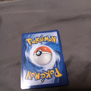 Puede incluir: Una carta coleccionable Pokémon azul con el logotipo de Pokémon y un diseño de Poké Ball. La tarjeta tiene una Poké Ball blanca y roja en el centro, sobre un fondo azul arremolinado. La palabra "Pokémon" está impresa en amarillo.
