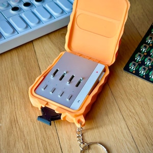 May include: A portable electronic music synthesizer housed in a bright orange protective case with a hinged lid. The device features a silver faceplate with four vertical sliders and a small red triangle icon. A metal keychain ring is attached to the base.