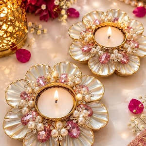 May include: Two flower-shaped candle holders featuring white shell-like petals, gold-toned metal centres, and decorative accents of small white pearls and oval pink crystals. Each holder contains a lit white tea light candle with a visible flame.