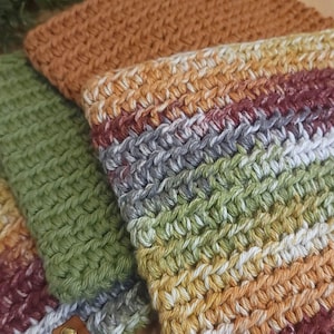 May include: A collection of stacked crochet pouches featuring textured yarn in various colors including solid rust orange, solid sage green, and a variegated pattern of white, gray, sage green, rust orange, and deep burgundy stripes.