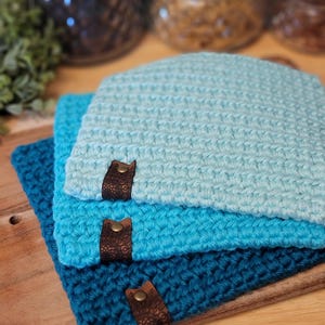 May include: Three crocheted beanies in shades of blue, stacked on a wooden tray. Each beanie has a brown leather tag with a decorative pattern and a metal rivet. The beanies are handmade and have a textured stitch.