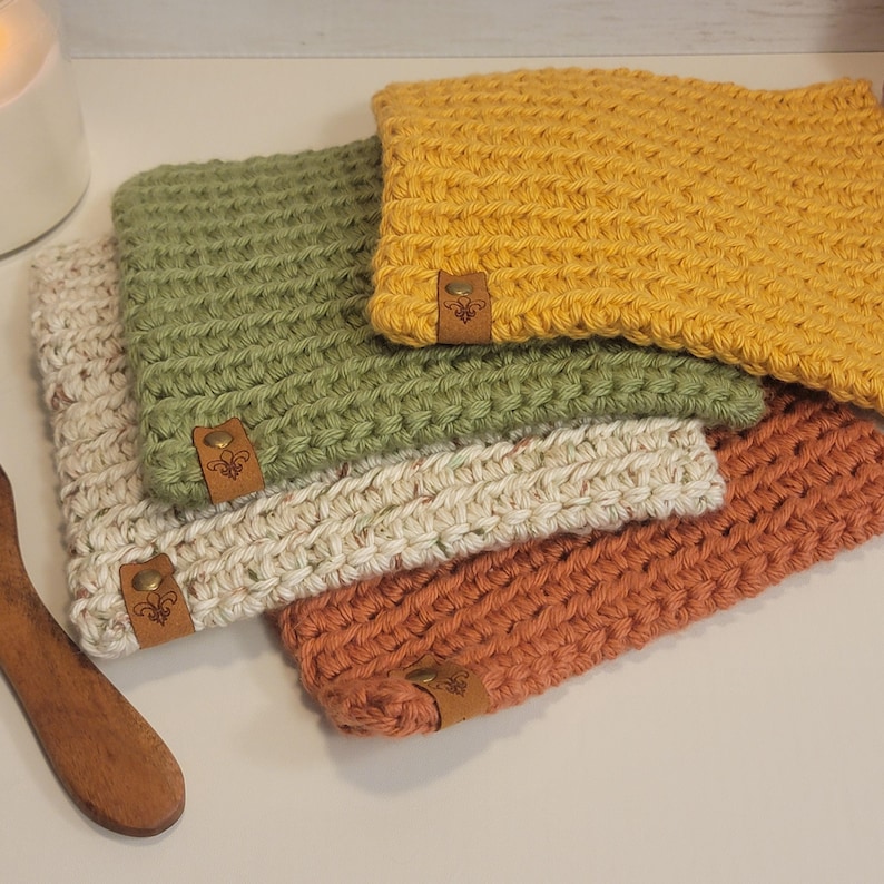 May include: A stack of five handmade crochet coasters in various colors: cream, olive green, mustard yellow, and burnt orange. Each coaster has a textured stitch pattern and a small, decorative leather tag with a fleur-de-lis design.