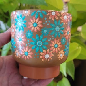 May include: A brown cylindrical planter featuring a hand-painted floral pattern with alternating teal and peach-colored petals, accented by small white and teal dots scattered across the surface, with a solid terracotta-colored base.