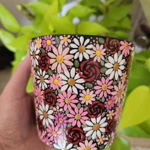 May include: A black ceramic flower pot with a hand-painted floral design. The pot is covered in white, pink, and yellow daisies, and red roses. The pot is cylindrical and has a glossy finish.