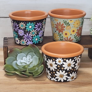 May include: Three terracotta plant pots with painted floral designs. One pot features purple and teal flowers on black, one has yellow and orange flowers on light green, and one displays white daisies on black. A faux succulent is placed nearby.
