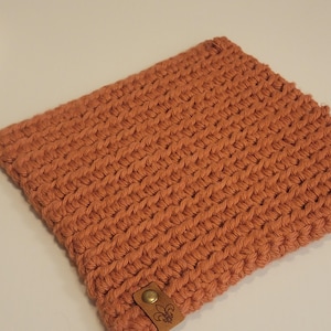 May include: A rectangular, hand-crocheted item in a warm, terracotta color. The textured surface features a tight, woven pattern. A small, brown leather tag with a brass button and a fleur-de-lis emblem is attached to the bottom.