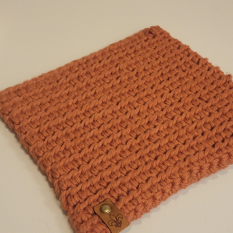 May include: A rectangular, hand-crocheted item in a warm, terracotta color. The textured surface features a tight, woven pattern. A small, brown leather tag with a brass button and a fleur-de-lis emblem is attached to the bottom.