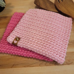 May include: Two handmade, pink crochet potholders. One is a darker shade of pink, and the other is a lighter shade. The lighter pink potholder has a small, brown leather tag with a heart-shaped detail. Wooden cooking utensils are in the background.