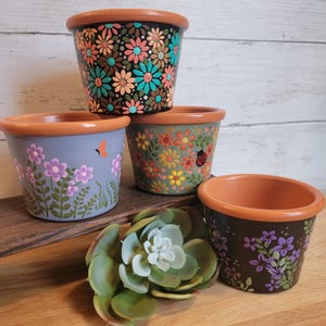 May include: Four hand-painted terracotta flower pots with floral designs. The pots feature a variety of colors, including black, blue, green, and purple, with orange rims. A succulent plant sits in front of the pots.