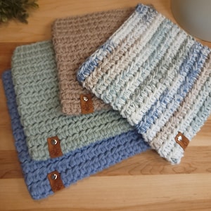 May include: Four crocheted pot holders in blue, green, beige, and blue and white stripes. Each pot holder has a brown leather tag with a fleur-de-lis design.