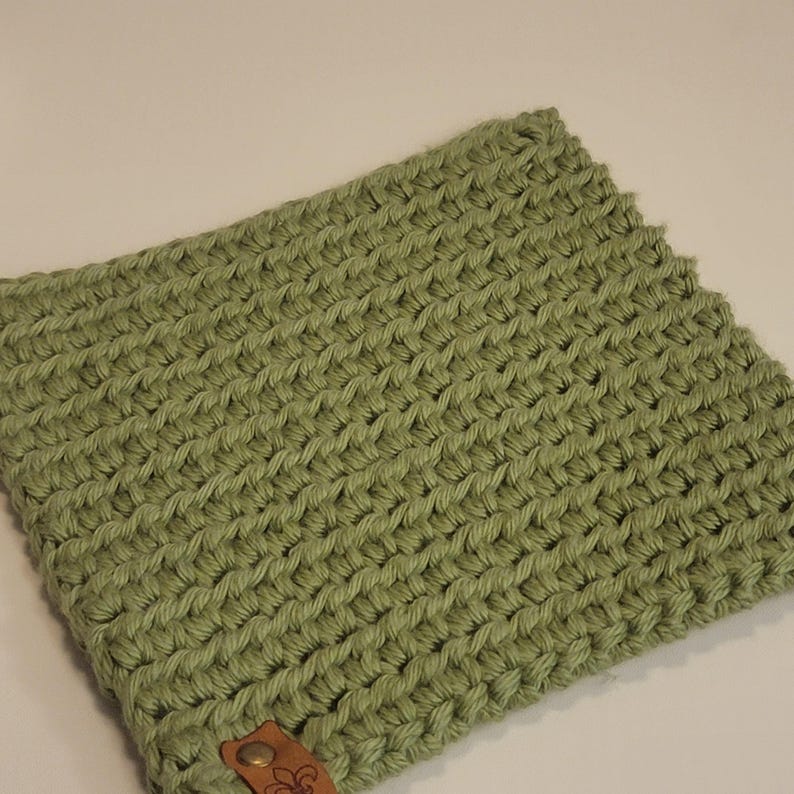 May include: A sage green, hand-crocheted cowl scarf. The scarf features a textured stitch pattern and a small, brown leather tag with a decorative emblem. The cowl is made of a soft, thick yarn.