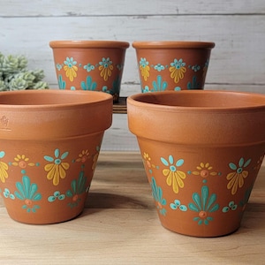 May include: Four terracotta flower pots arranged in two rows. Each pot features a hand-painted pattern of yellow and teal floral motifs encircling the exterior. The pots have a wide rim and a tapered base, with a small stamped logo on the side.