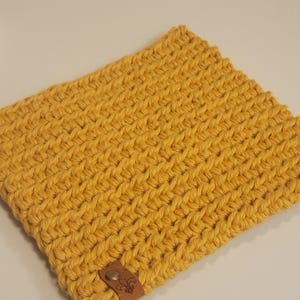 May include: A square, hand-crocheted item in a vibrant yellow hue. The textured surface features a repeating pattern of raised stitches. A small, brown leather tag with a decorative emblem is attached to the corner.