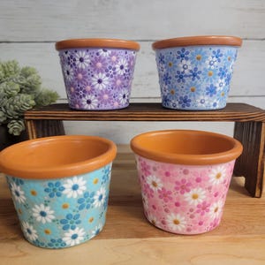 May include: Four small, ceramic flower pots with a terracotta rim. The pots are decorated with a daisy pattern in shades of purple, blue, turquoise, and pink. The pots are displayed on a small wooden shelf.