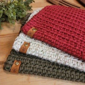 May include: Three crocheted pot holders in red, white, and green with brown leather labels. Each label has a fleur-de-lis design.