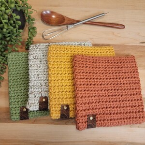 May include: A collection of four square, crocheted washcloths in shades of green, gray, yellow, and orange. Each cloth has a leather tag with a decorative pattern. A wooden spoon and whisk rest above the cloths.